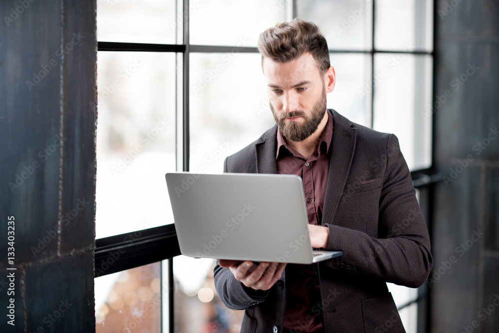 Handsome bearded businessman dressed in the suit standing with laptop near the big window in the lof