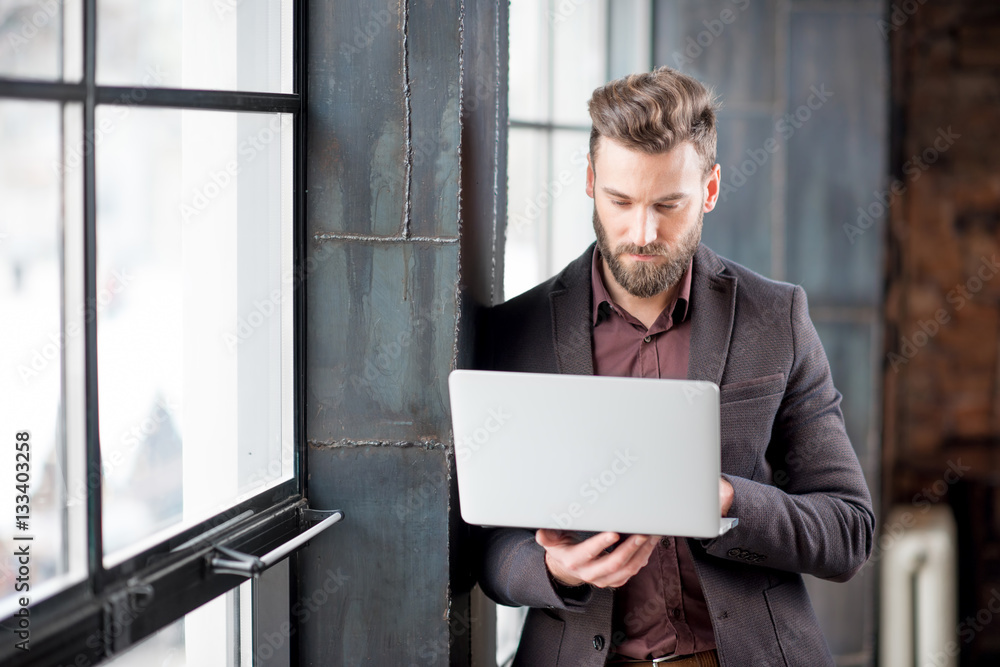 Handsome bearded businessman dressed in the suit standing with laptop near the big window in the lof