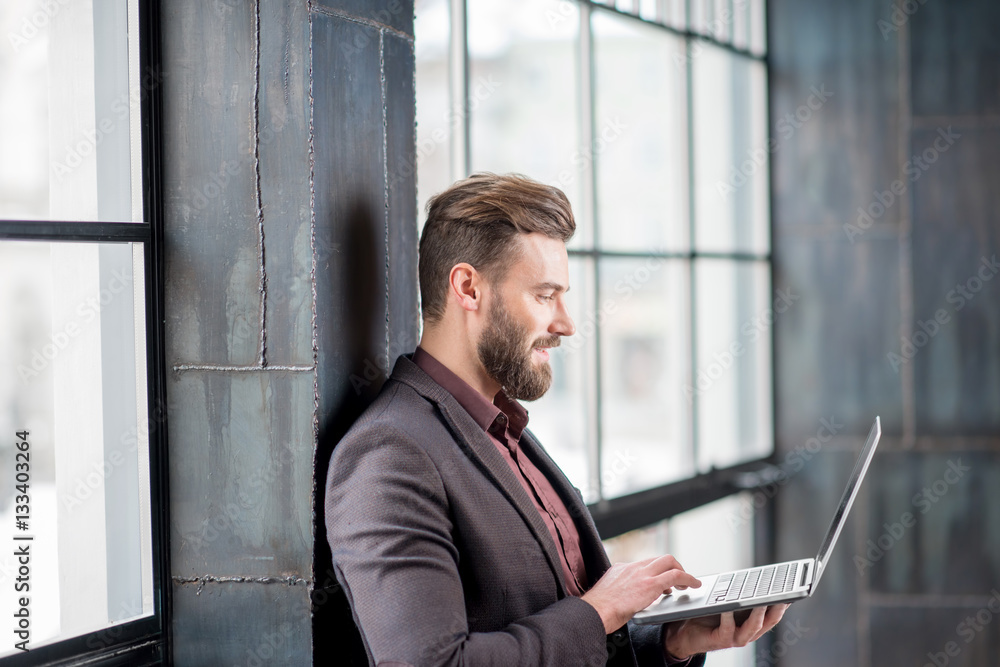 Handsome bearded businessman dressed in the suit standing with laptop near the big window in the lof