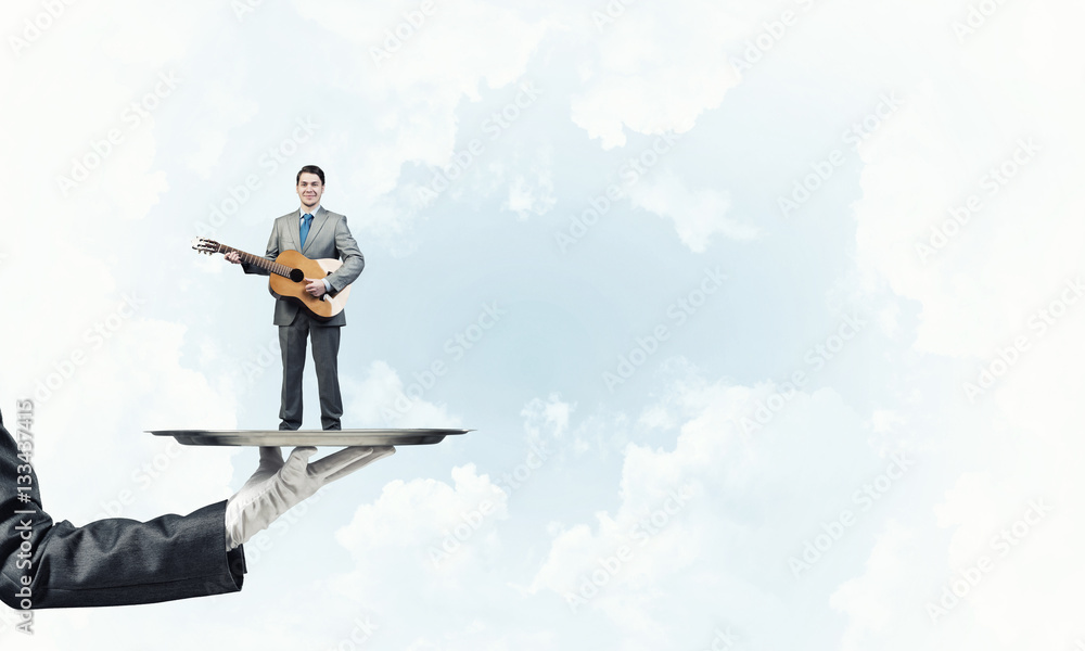 Businessman on metal tray playing acoustic guitar against blue sky background