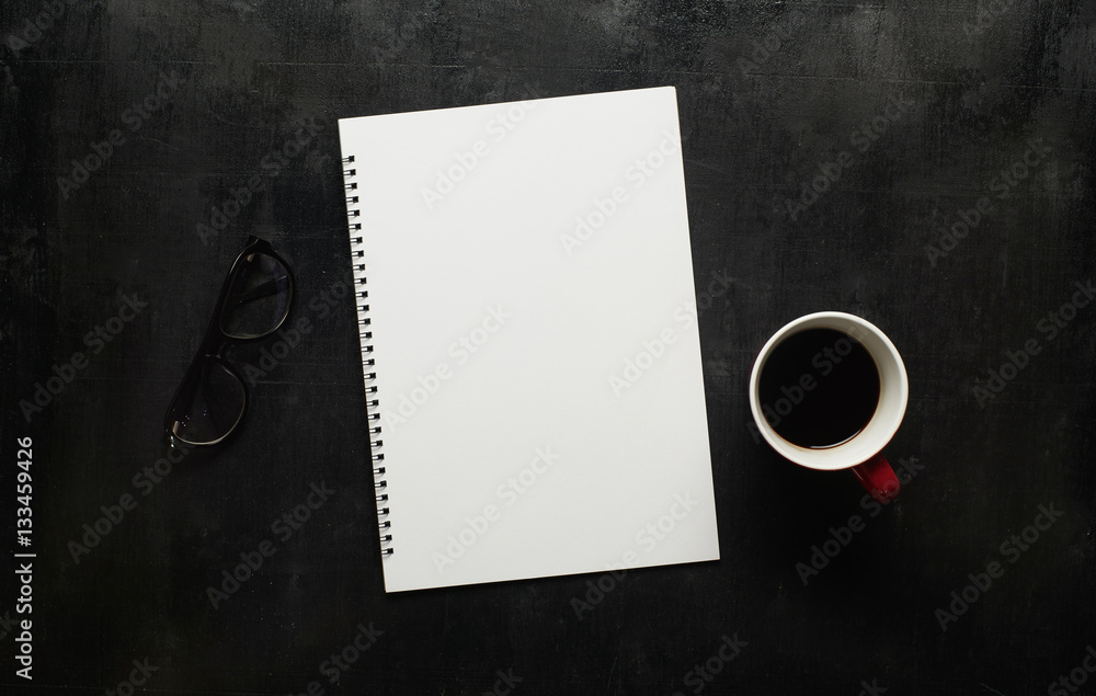 Wooden black office desk table with notebook, eyeglasses and  cup of coffee. Blank notebook page for