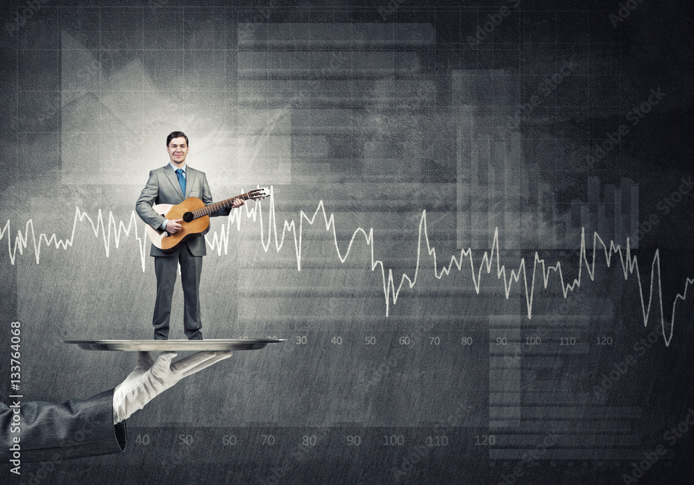 Businessman on metal tray playing acoustic guitar against concrete wall background with charts