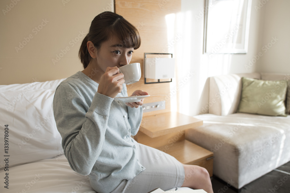 The woman is sitting in the bed in the hotel room and drinking coffee