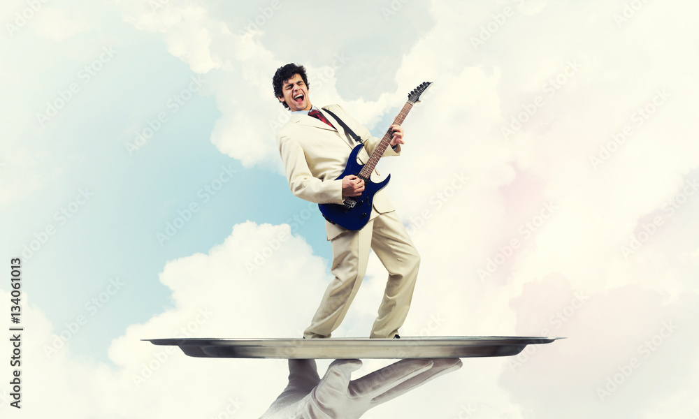 Businessman on metal tray playing electric guitar against blue sky background