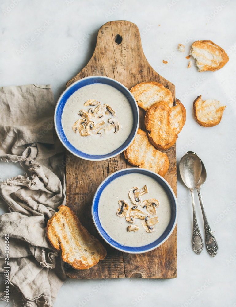 Creamy mushroom soup in bowls with toasted bread slices on rustic serving board over grey marble bac