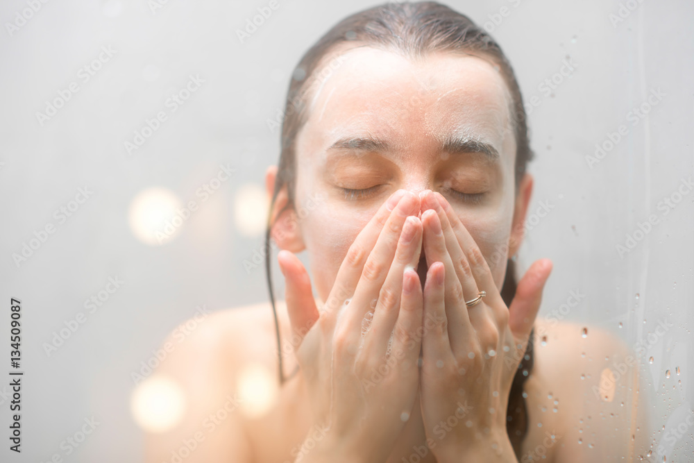 Close-up portrait of a woman with soap on her wet face standing behind the glass in the shower. Imag