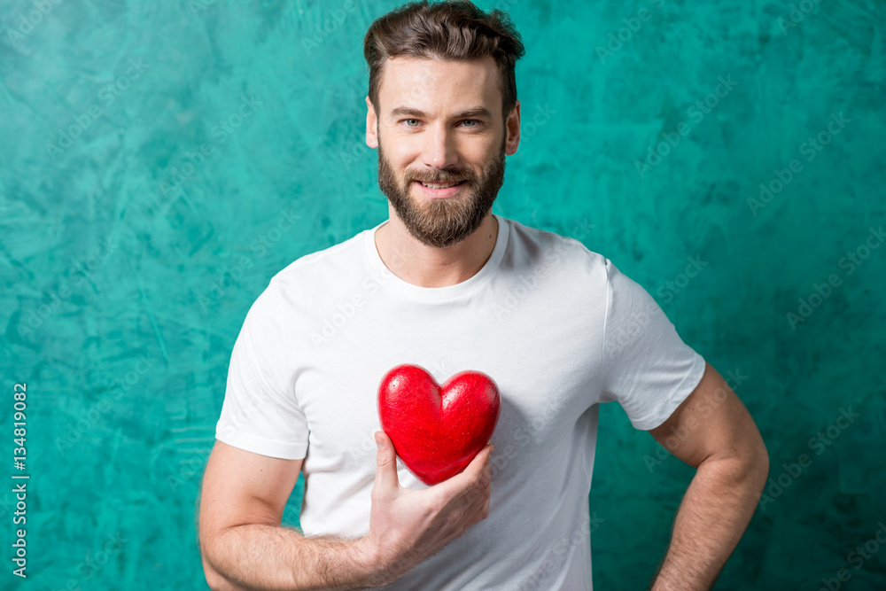 Handsome man in the white t-shirt holding red heart on the painted green wall background. Valentine