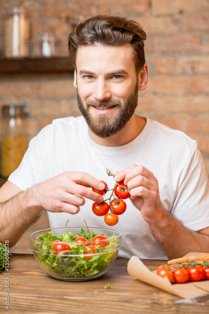 Handsome bearded man in white t-shirt making salad with tomatoes and pepper in the kitchen. Healthy 