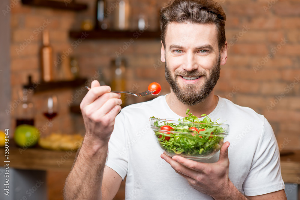 Handsome bearded man in white t-shirt eating salad with tomatoes in the kitchen. Healthy and vegan f