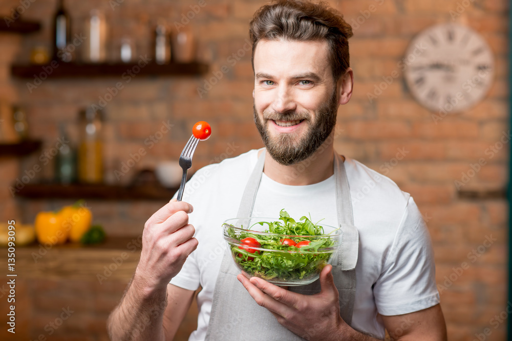 Handsome bearded man in white t-shirt and apron eating salad with tomatoes in the kitchen. Healthy a