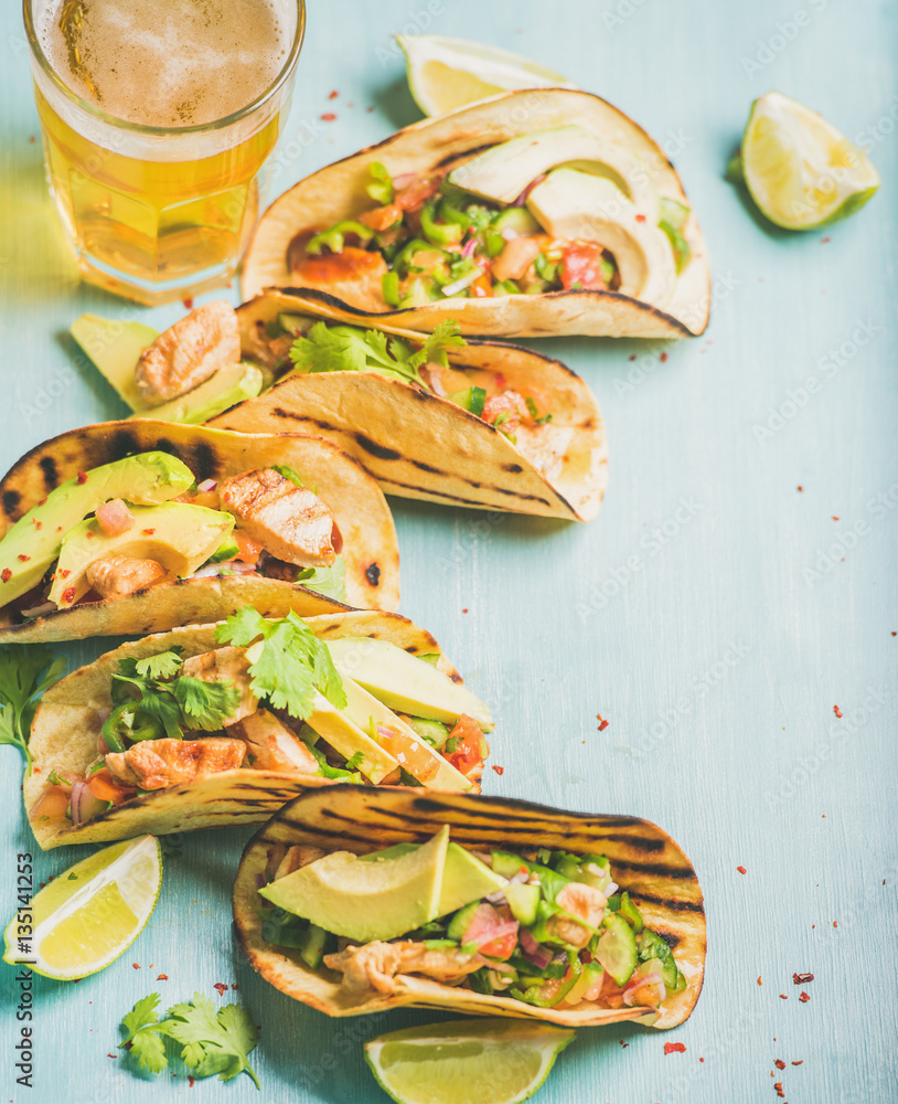 Healthy corn tortillas with grilled chicken, avocado, fresh salsa, limes and beer in glass over blue