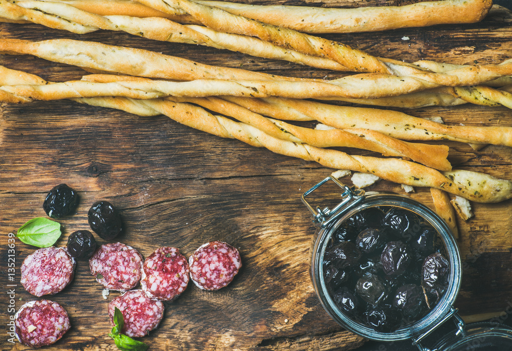 Close-up of Italian Grissini bread sticks heap, dry cured pork meat sausage slices and black olives 