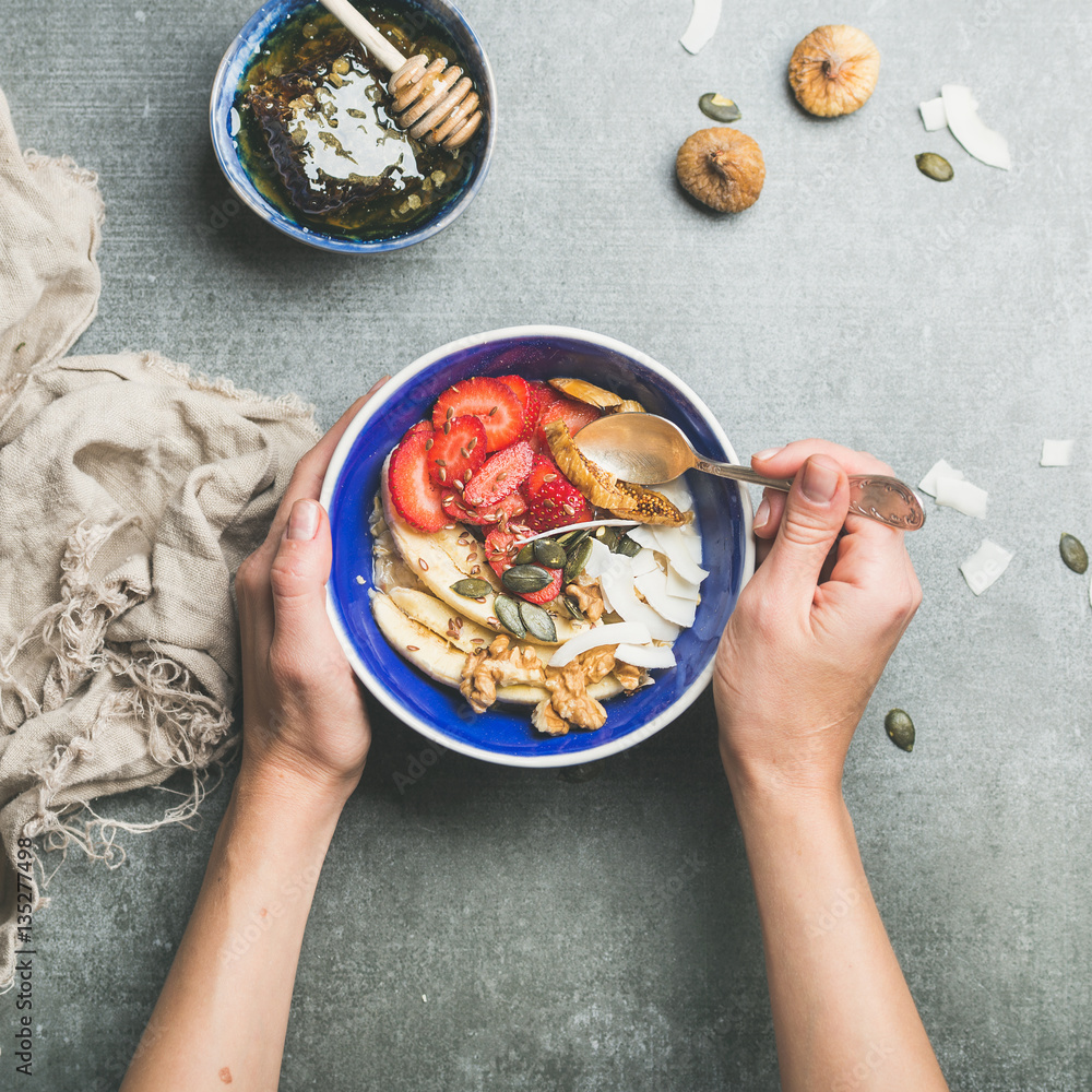 Yogurt, granola, seeds, fresh and dry fruit and honey in blue ceramic bowl in woman s hands over gr