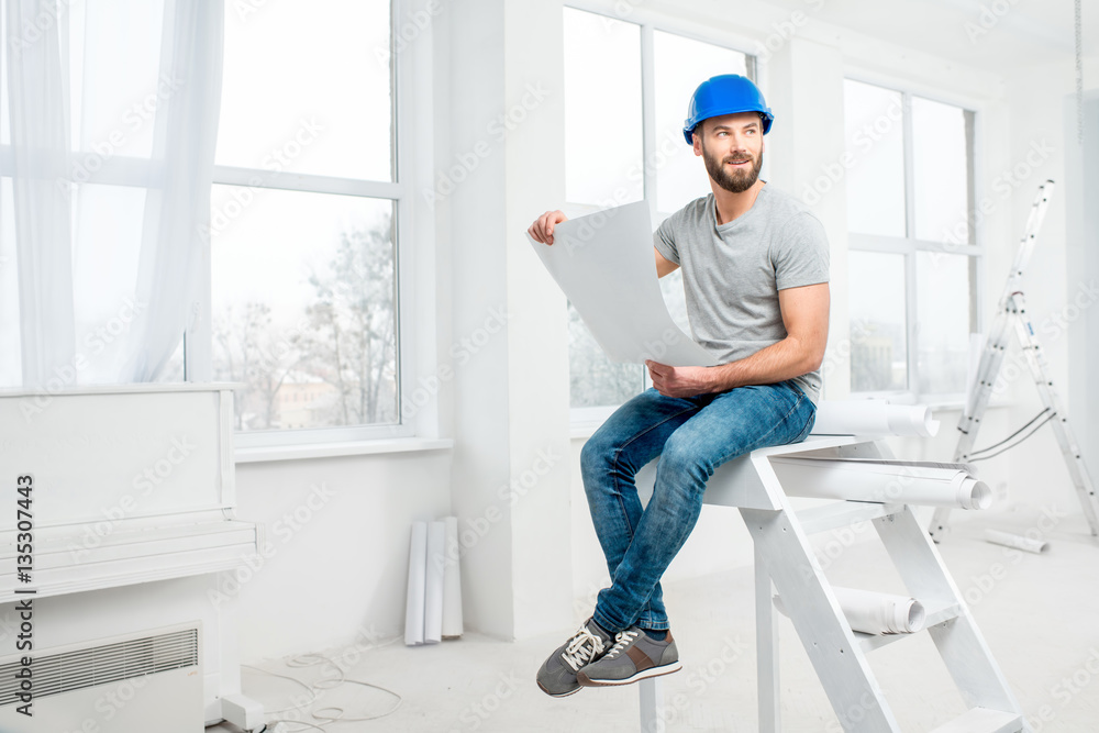 Handsome repairman or builder in helmet working with drawings on renovation of apartment interior