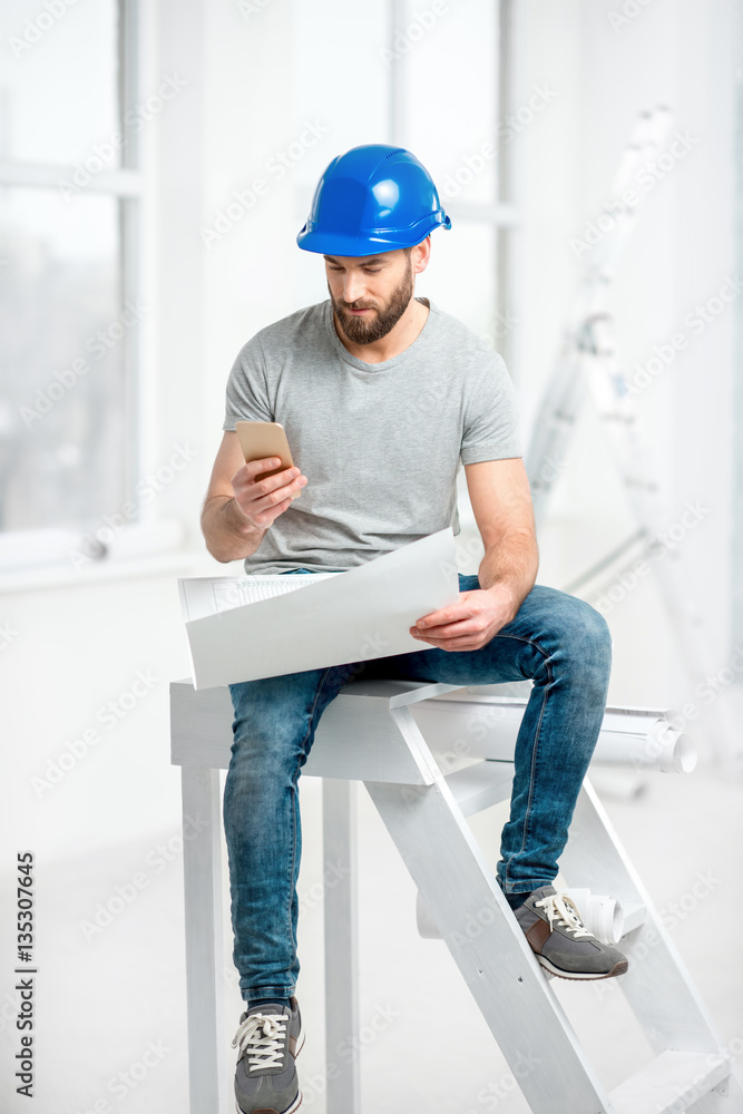 Handsome repairman or builder in helmet working with drawings and phone on the renovation of apartme