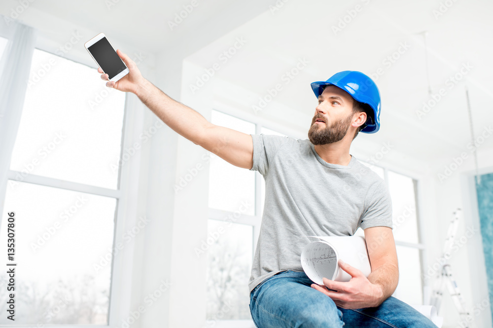 Handsome repairman or builder in helmet with phone and drawings in the white interior