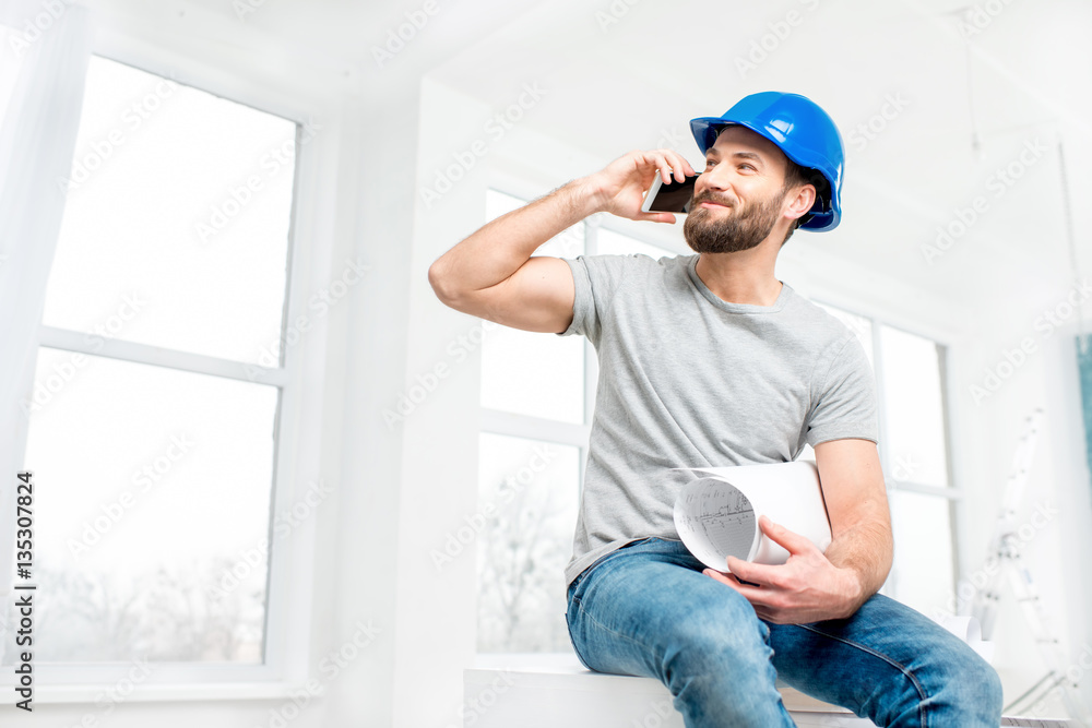 Handsome repairman or builder in helmet talking with phone with drawings in the white interior