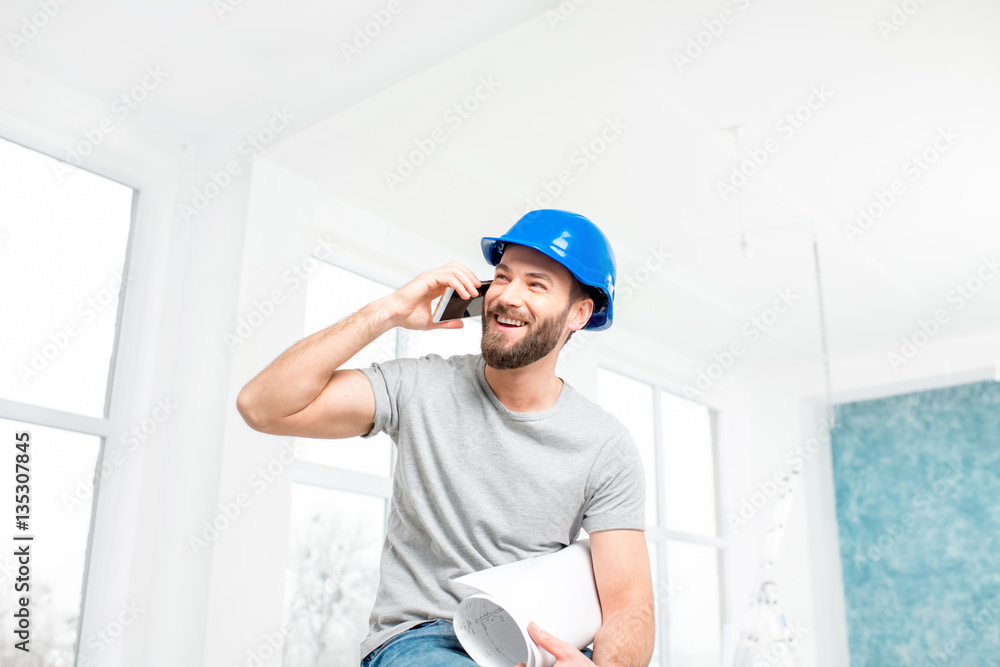 Handsome smiling repairman or builder in helmet talking with phone with drawings in the white interi