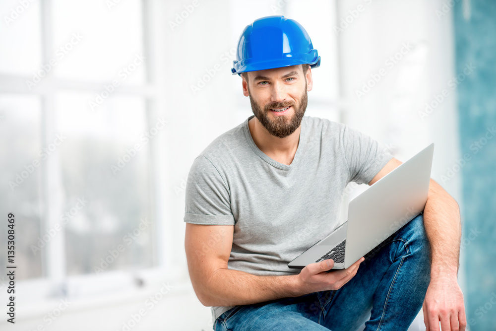 Portrait of a handsome builder, foreman or repairman in the helmet sitting with laptop in the white 