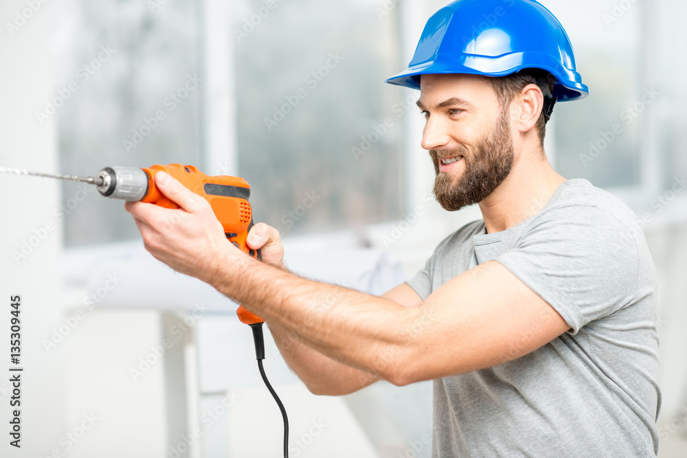 Handsome repairman in protective helmet drilling the wall indoors