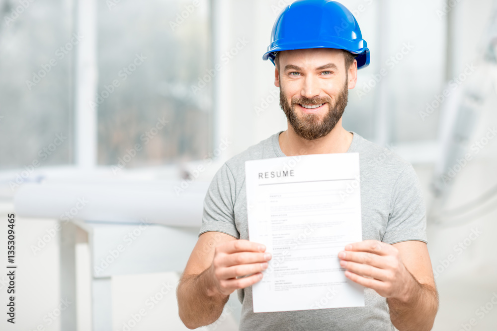 Handsome builder or worker in protective helmet holding his resume standing in the white interior.