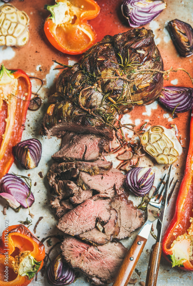 Cooked Roastbeef meat with roasted vegetables and herbs in metal baking tray, top view, vertical com