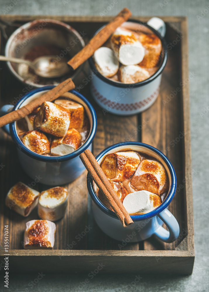 Hot chocolate in enamel mugs with cinnamon and roasted marshmallows in wooden tray over grey backgro