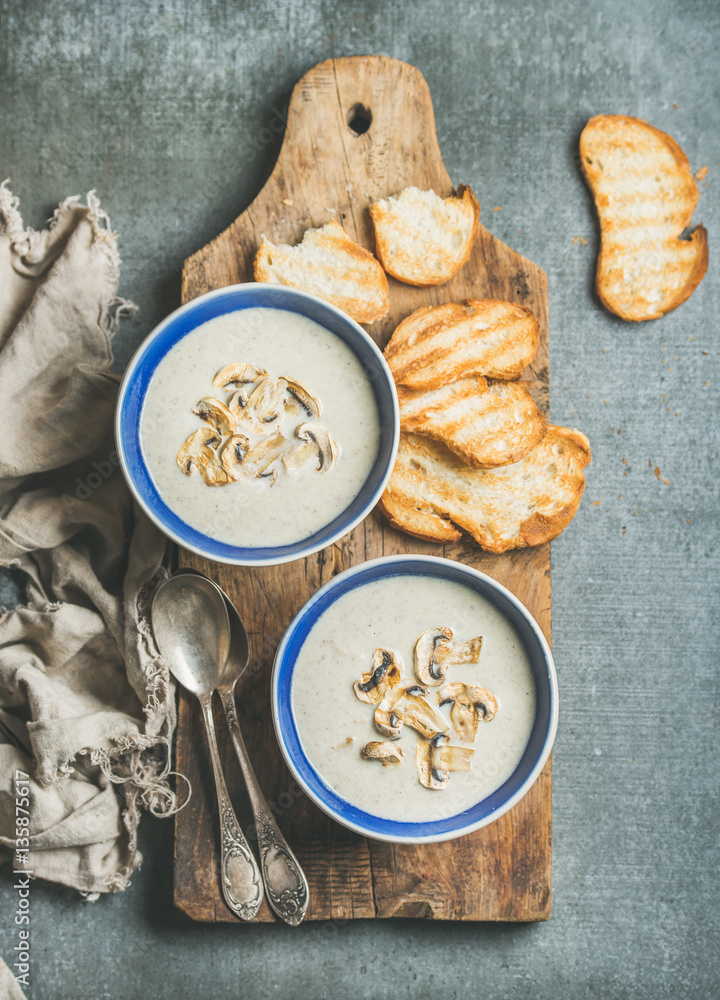 Creamy mushroom soup in bowls with toasted bread slices on rustic serving board over grey concrete b