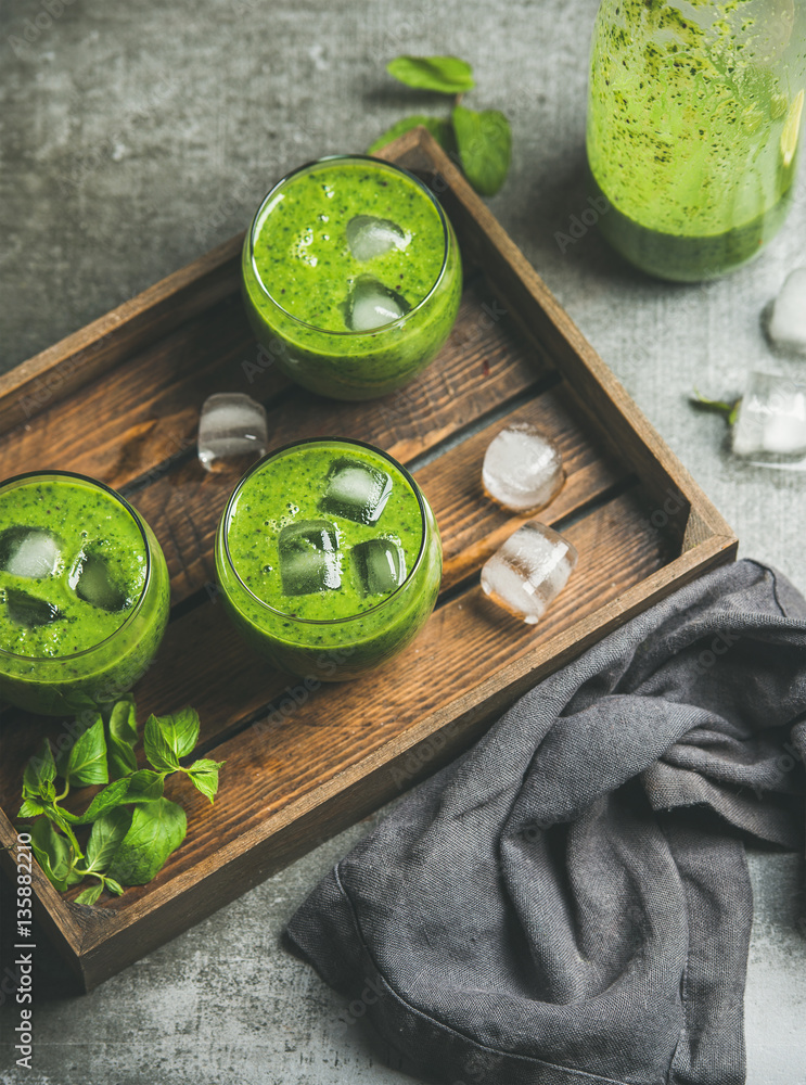 Fresh green smoothie in glasses with ice cubes, mint and lime in wooden tray over grey concrete back