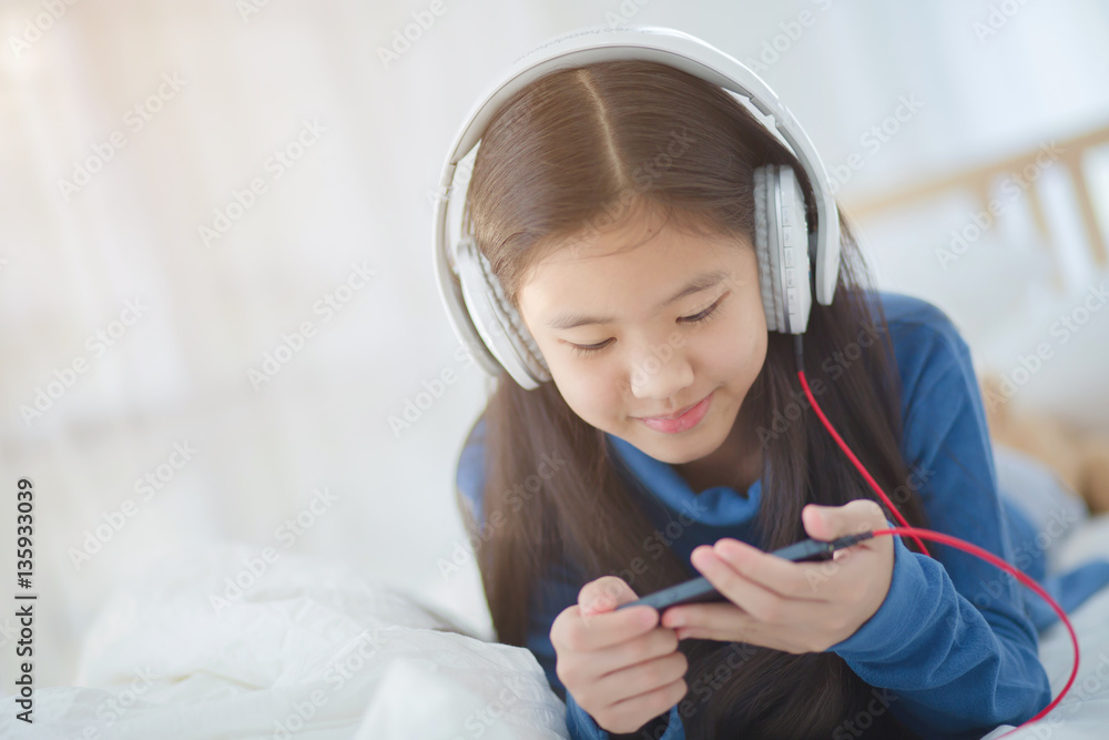 Pretty Asian girl using headphone for listen music by smartphone on the bed in her bedroom