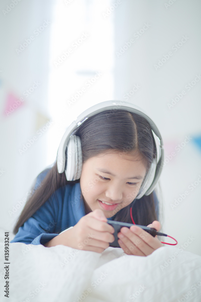 Pretty Asian girl using headphone for listen music by smartphone on the bed in her decorated bedroom