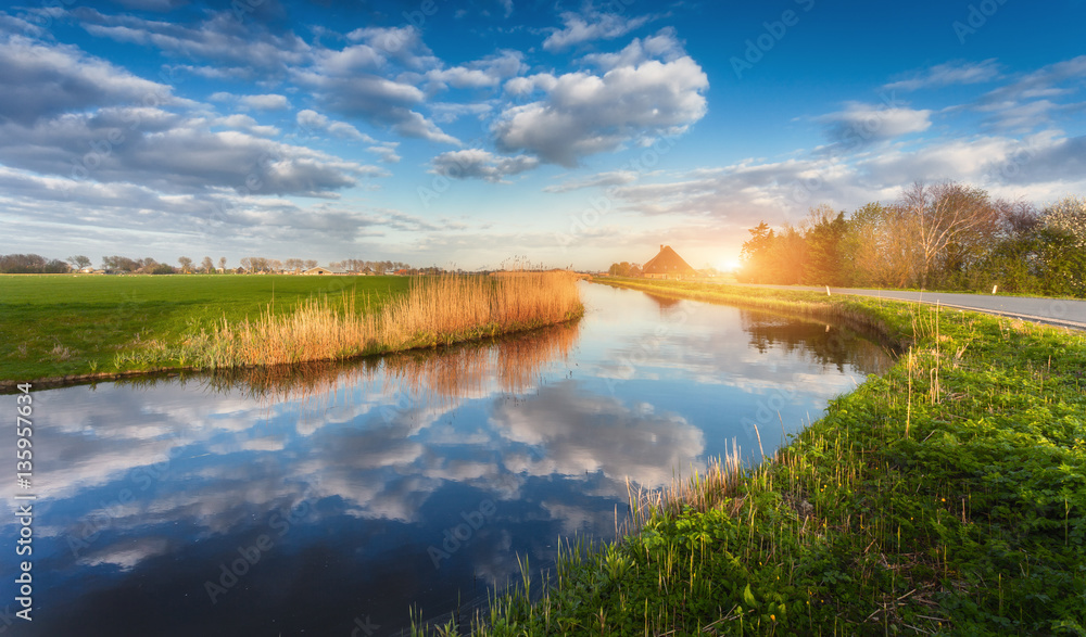 Buildings and trees near the water canal at sunrise in Netherlands. Colorful blue sky with clouds. S