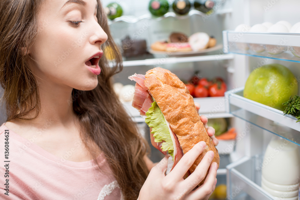 Young woman eating big sandwich in front of the refrigerator full of friuts and vegetables