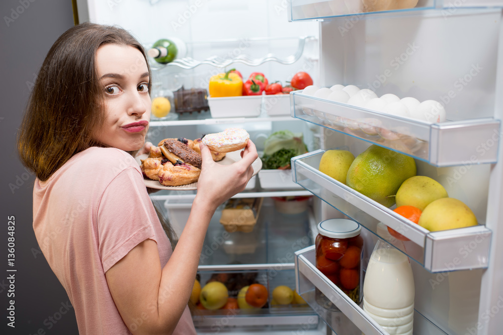 Young woman in the sleepwear eating sweet donuts near the refrigerator