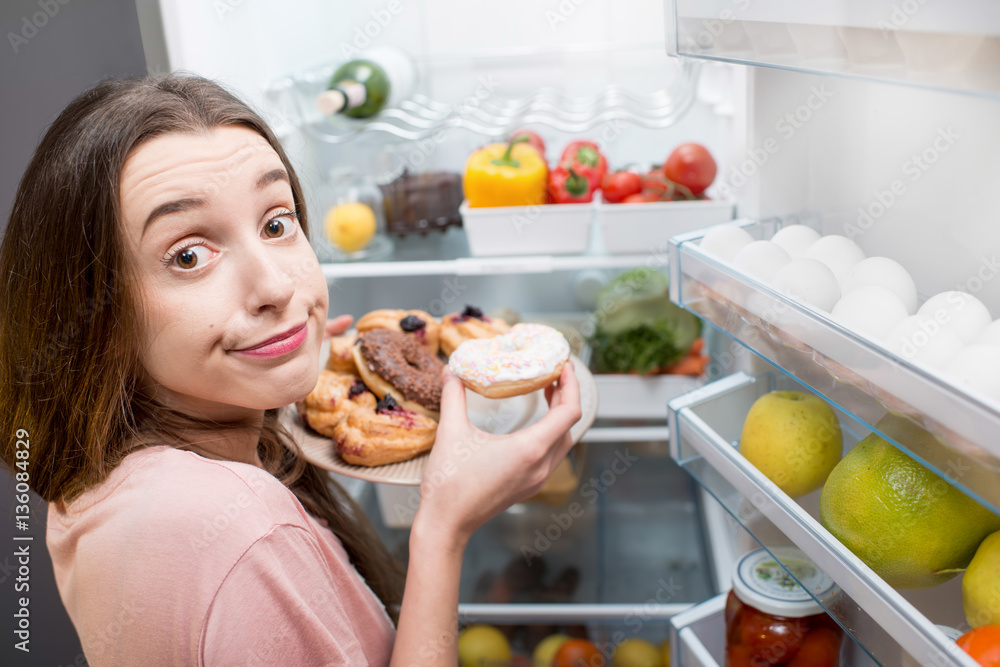 Young woman in the sleepwear eating sweet donuts near the refrigerator