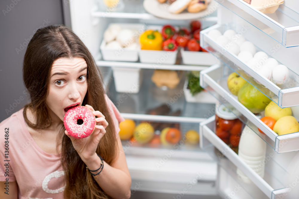 Portrait of a young sad woman in the sleepwear with sweet donuts standing near the open refrigerator