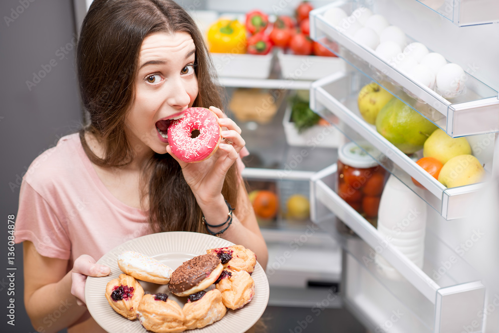 Young woman in the sleepwear eating sweet donuts near the refrigerator