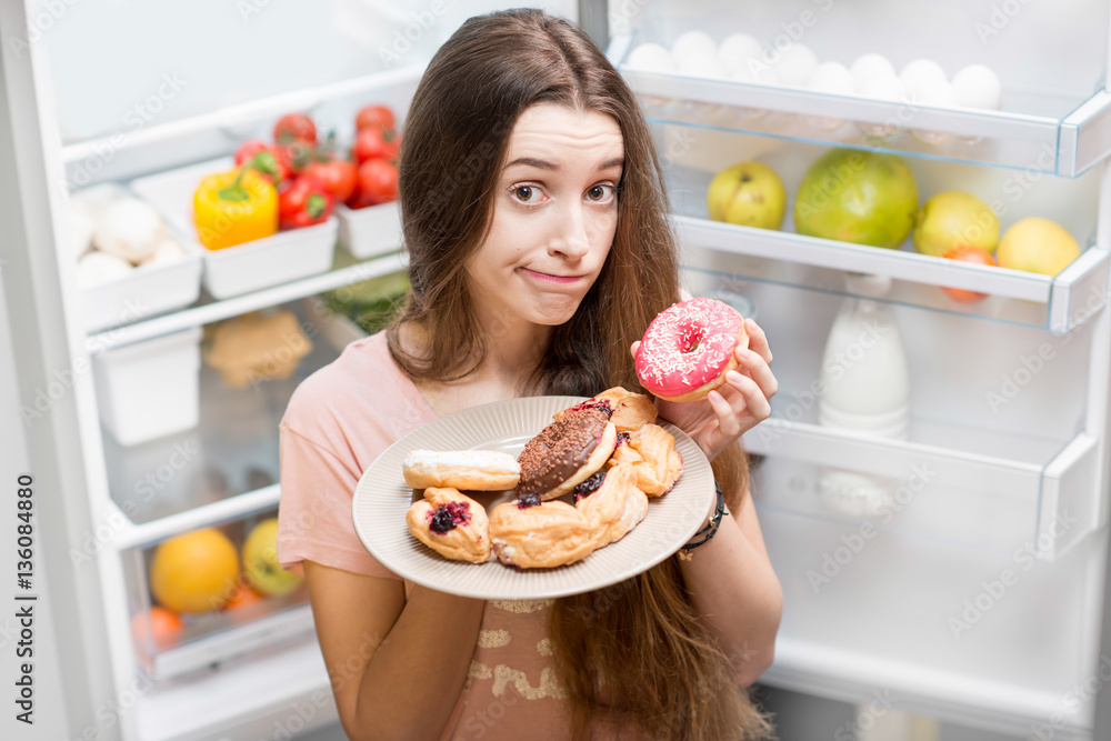 Portrait of a young sad woman in the sleepwear with sweet donuts standing near the open refrigerator