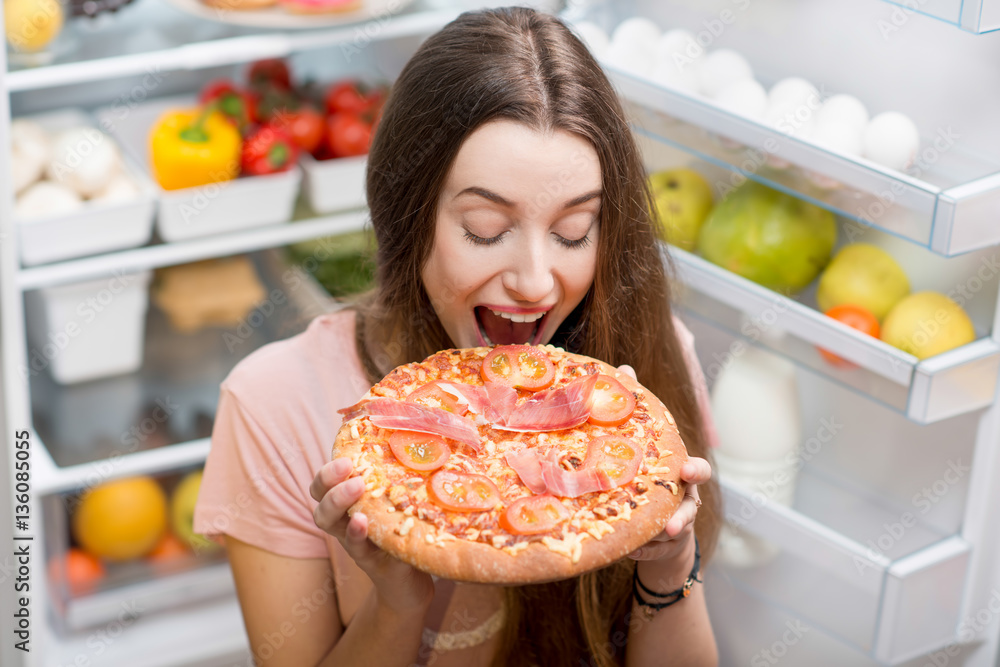 Young woman with pizza standing near the open refrigerator full of fruits and vegetales