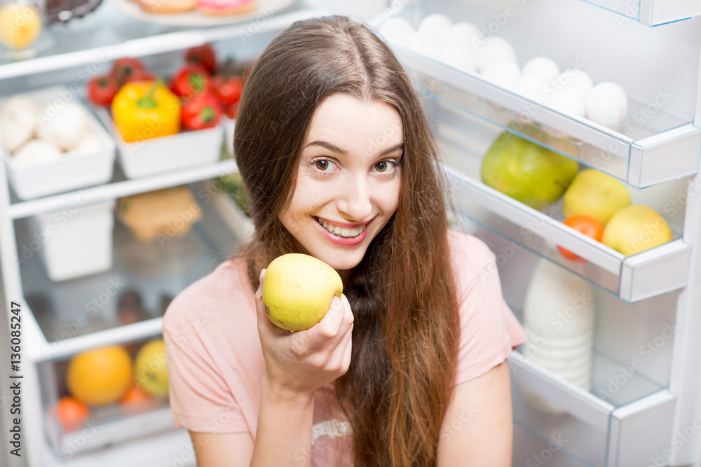 Portrait of a young smiling woman with apple standing in front of the open refrigerator