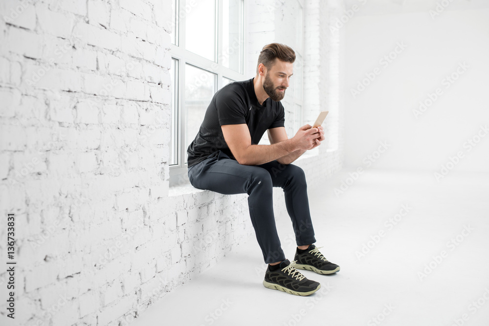 Handsome man in the black sportswear sitting with phone on the window in the white gym interior