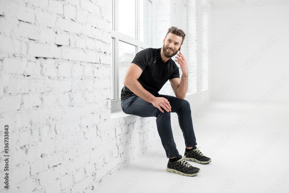 Handsome man in the black sportswear talking with phone sitting on the window in the white gym inter