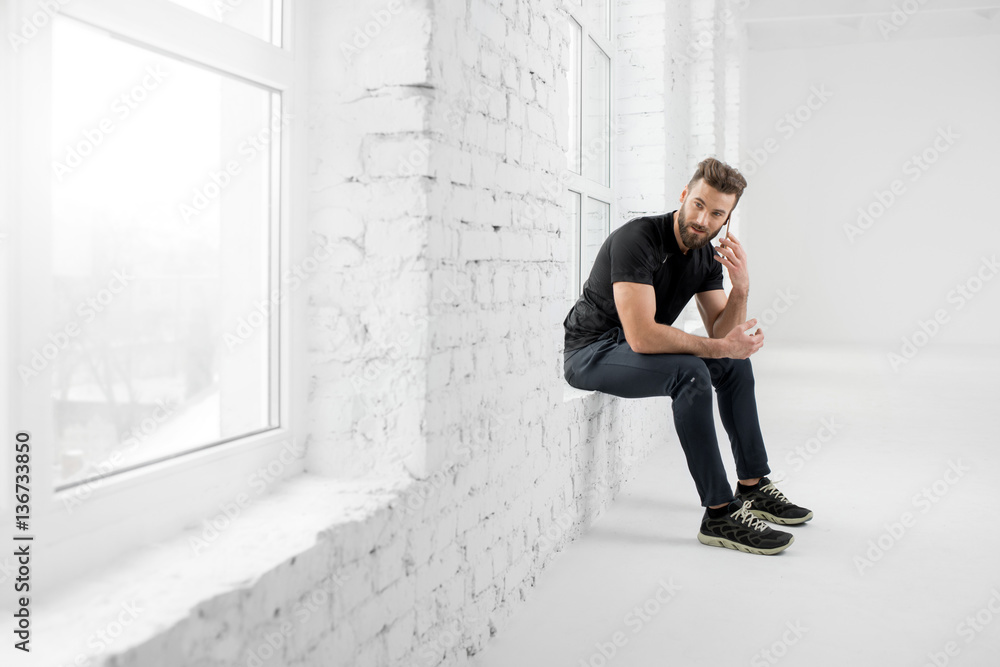 Handsome man in the black sportswear talking with phone sitting on the window in the white gym inter