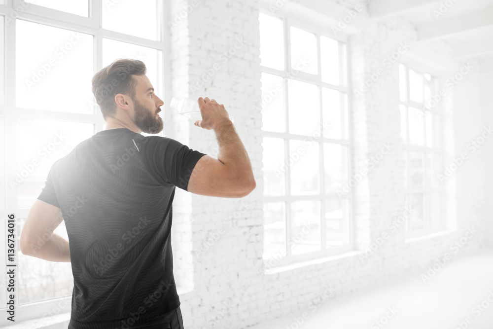 Man in the black sportswear drinking water in the white gym interior