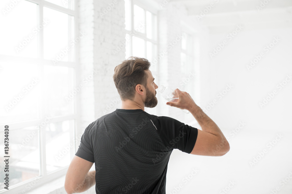 Man in the black sportswear drinking water in the white gym interior