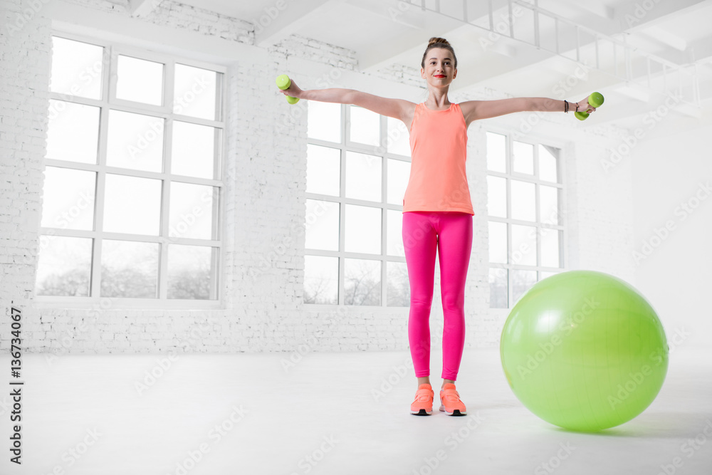 Young woman in colorful sportswear lifting dumbbels in the white gym with green fitness ball on the 