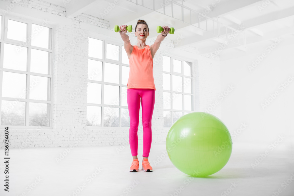 Young woman in colorful sportswear lifting dumbbels in the white gym with green fitness ball on the 