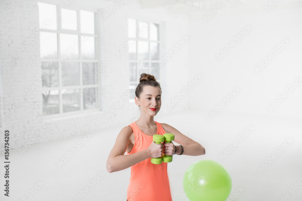 Young woman in colorful sportswear lifting dumbbels in the white gym with green fitness ball on the 