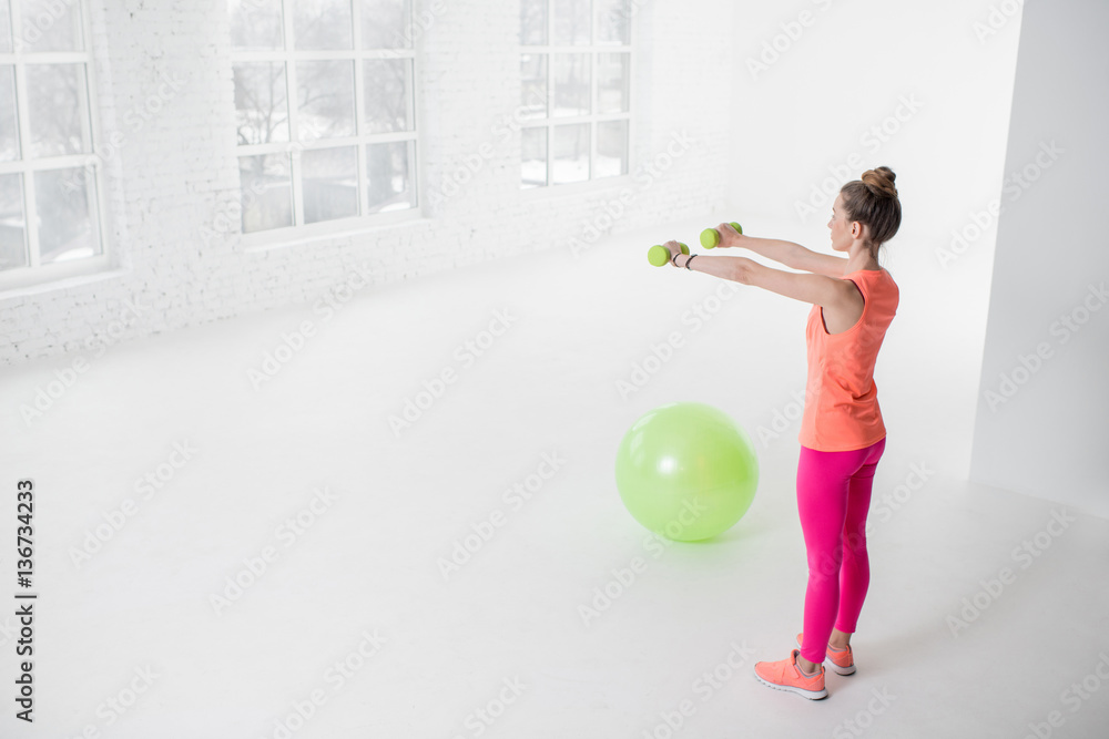 Young woman in colorful sportswear lifting dumbbels in the white room with green fitness ball on the