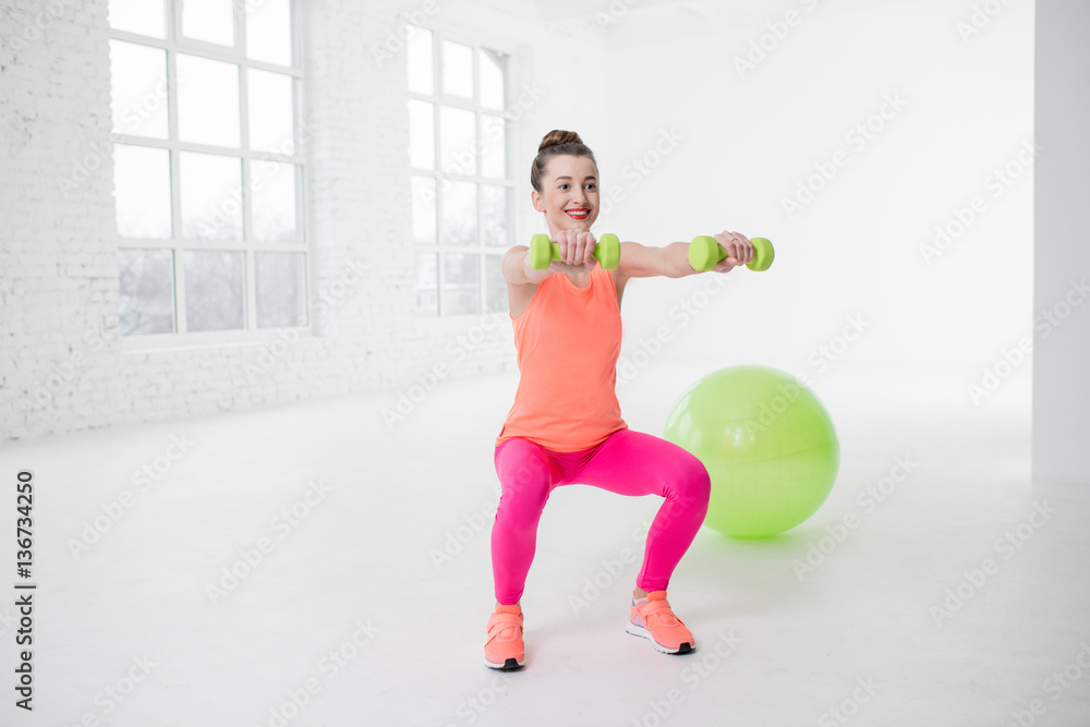 Young woman in colorful sportswear squatting with dumbbels in the white gym with fitness ball on the